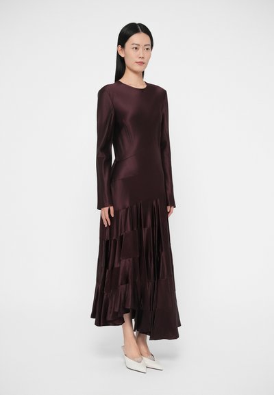 Burgundska satenasta obleka z dolgimi rokavi in zloženo volanasto spodnjico, ki ima prilegajoči se bodi in okrogel izrez. Model nosi bele čevlje z ostrim prstom.