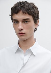 White button-up shirt with a classic collar, featuring a smooth texture and a slim fit. Model has short, dark hair and light skin.