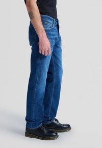 Mężczyzna ubrany w niebieskie dżinsy, czarne buty i czarną koszulkę stojący bokiem na jednolitym jasnoszarym tle.