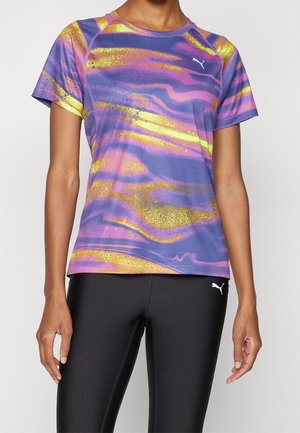 Personne portant un maillot de sport à manches courtes avec un motif abstrait violet, jaune et rose et des leggings noirs avec des logos Puma.
