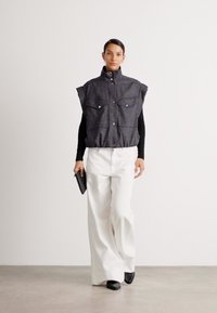 Grå ärmlös vest med to frontlommer, båret over en svart topp, kombinert med hvite vide bukser og svarte sko. Minimalistisk design.
