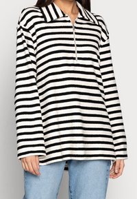 Gestreifter Pullover in Weiß und Schwarz, mit einem halben Reißverschluss, Kragen, langen Ärmeln und einer weichen Textur. Lässiger Schnitt.