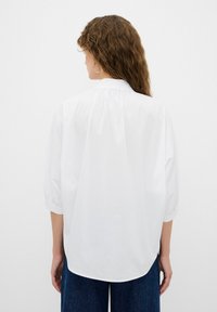 Hvid skjorte med korte ærmer, der har en afslappet pasform, plisseret rygdetalje og rundet kant, parret med mørkeblå, løs siddende jeans.