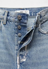 Spijkerbroek met een lichtblauwe wassing, voorzien van een knoopsluiting, metalen hardware en zichtbare stiksels. Verweerd textuur nabij de zakken.