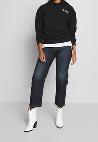 Sudadera negra con capucha y gráfico frontal, llevada sobre una camisa blanca, combinada con jeans azul oscuro y botas blancas hasta el tobillo con tacón cuadrado.
