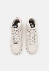 Baskets Nike Air Force 1 en cuir beige clair, avec des accents rouges, une boîte à orteils texturée, des lacets blancs et un col rembourré.
