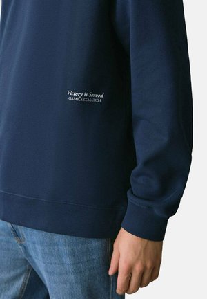 Felpa blu navy con scollatura a girocollo, polsini e orlo inferiori a coste. Presenta la scritta bianca "Victory is Served GAME, SET, MATCH" sul lato.