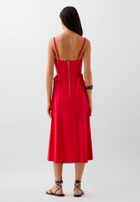 Robe midi rouge sans manches avec un dos droit, fermeture éclair et détails noués à la taille. Texture lisse et silhouette fluide.