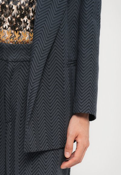 Blazer scuro a motivo spina di pesce con una trama strutturata e una vestibilità su misura. Include una tasca frontale, visibile parzialmente sul lato sinistro.