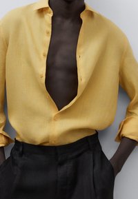 Camisa de lino amarilla con un cuello abierto y varios botones visibles. Combinada con pantalones negros, que muestran un corte holgado y una textura suave.