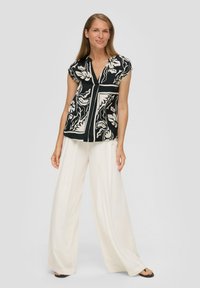 Schwarzes Oberteil mit weißem Blumenmuster, kurzen Ärmeln und V-Ausschnitt; kombiniert mitweiten weißen Hosen und schwarzen Sandalen. Glatte Stofftextur.