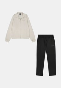 Chaqueta cremoso claro con cierre y cuello alto, con un logo bordado, combinada con pantalones de chándal negros con cintura elástica y logo.