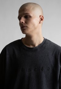 Νεαρός με ξυρισμένο κεφάλι, που φοράει σκουρόχρωμο ξεθωριασμένο μπλουζάκι, κοιτάζει προς το πλάι μπροστά σε απλό γκρι φόντο.