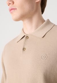 Bēša adīta polo krekla ar mazu izšūtu logotipu, divām pogām ap kaklu un mīkstu, teksturētu audumu.