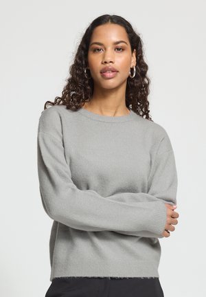 Jeune femme aux cheveux bouclés porte un pull gris à manches longues et des boucles d’oreilles argentées, debout, bras croisés devant un fond uni.