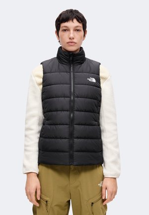 Person trägt eine schwarze gesteppte Weste über einer weißen Fleecejacke und olivgrüne Hose mit Reißverschlusstaschen, steht vor einem einfarbigen Hintergrund.