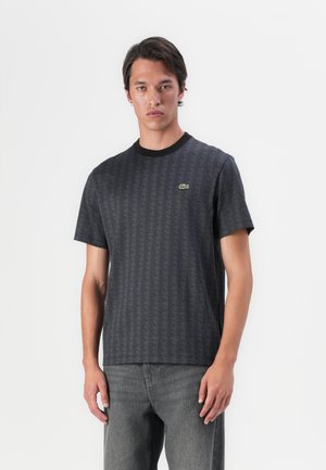 Tricou negru cu mânecă scurtă, cu un model geometric, având un guler rotund și un mic logo verde pe piept. Blugii gri completează ținuta.