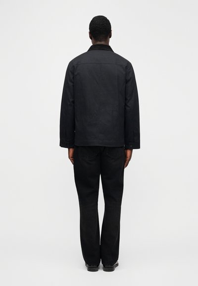 Μαύρο μπουφάν με  δομημένο γιακά, κλείσιμο με κουμπιά και μακριά μανίκια, φορεμένο πάνω από μαύρο τζιν με ίσια μπατζάκια, σε θέα από πίσω.