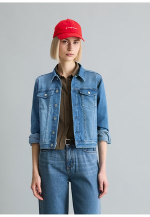 Frau mit kurzen blonden Haaren, die eine rote Mütze, eine blaue Jeansjacke mit hochgekrempelten Ärmeln, ein braunes Hemd mit Knöpfen und eine blaue Jeans vor grauem Hintergrund trägt.