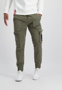 Pantalon cargo vert olive avec plusieurs poches, poignets élastiques, ceinture à cordon réglable et détails de patch noir.