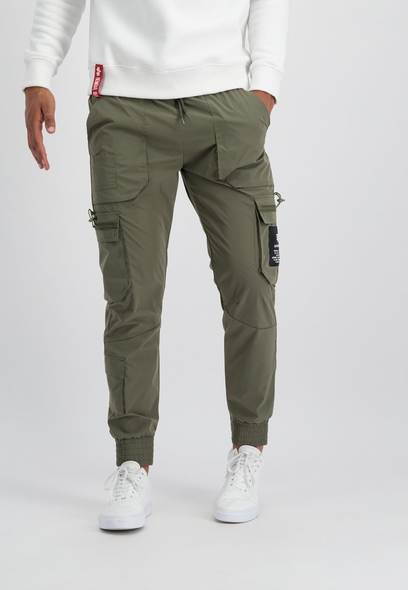 Pantalon cargo vert olive avec plusieurs poches, poignets élastiques, ceinture à cordon réglable et détails de patch noir.