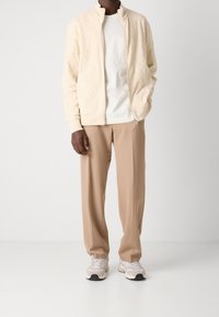 Man som bär en beige zip-up strukturerad jacka, vit T-shirt, beiga byxor och vita sneakers står mot en vit bakgrund.