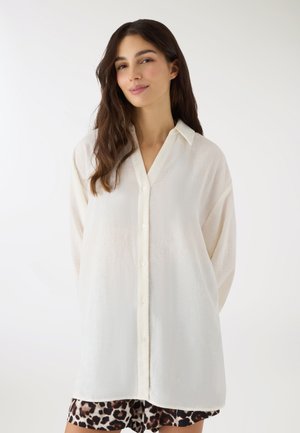 Giovane donna con lunghi capelli castani che indossa una camicia bianca oversize con bottoni e pantaloncini leopardati su sfondo bianco.