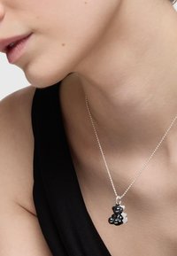Ciondolo a forma di orsetto nero su una catena d'argento, con una finitura lucida e una consistenza liscia, indossato sulla pelle nuda contro un top nero.