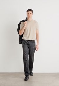 Beige kötött póló, sötét farmernadrágba tűrve. A modell egy fekete bőrkabátot tart a vállán. Fekete cipők. Egyszerű, modern dizájn.