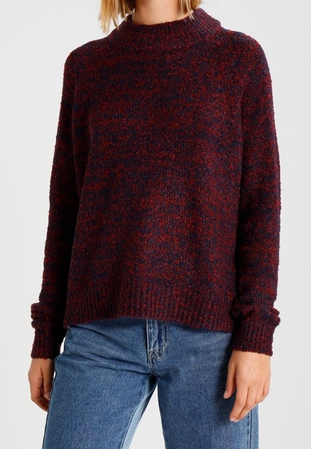 Personne portant un pull en maille texturée bordeaux et bleu marine à manches longues, accompagné d'un jean en denim bleu, se tenant devant un fond uni.