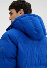 Niebieska kurtka puchowa z kapturem, z teksturowaną powierzchnią i wytłoczonym detalem logo. Wykonana z lekkiego, izolowanego materiału.