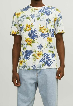 Mand iført en hvid kortærmet skjorte med gule, blå og grønne blomster- og palmemønstre, kombineret med lyseblå jeans.