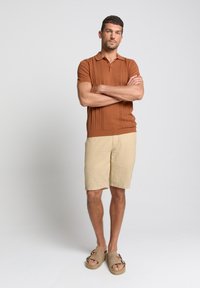 Bronzen gebreide poloshirt met textuurpatronen, gecombineerd met beige shorts. Gedragen met lichtbruine instap-sandalen. Neutrale achtergrond.