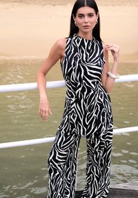 Mustvalge sebratriibuline kombinesoon laiade sääretega, varrukateta disain ja sile kangapind. Valge käevõruga aksentidega.