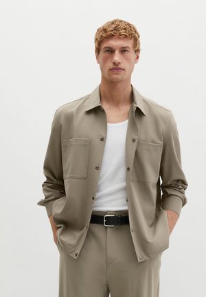 Homme blond portant une veste beige à boutons sur un débardeur blanc avec un pantalon beige assorti et une ceinture noire, mains dans les poches, fond neutre.