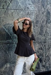 Frau in schwarzem Oberteil und weißen Hosen posiert vor grünem Marmorwand, trägt schwarze Sonnenbrille und hält eine grüne Clutch.