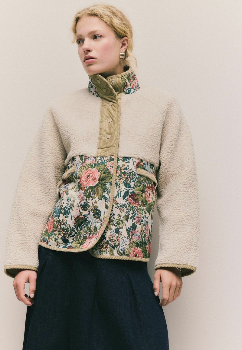 Fleece- og blomsterjakke med en beige overdel, en livlig blomstrende nederdel med pink blomster, matchende grønne kanter og trykknaplukning.