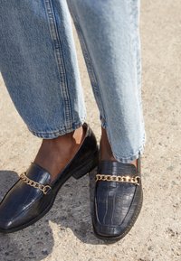 Mocassini neri in pelle con goffratura a coccodrillo e un accento di catena dorata, abbinati a jeans in denim azzurro chiaro, leggermente arrotolati, su una superficie testurizzata.