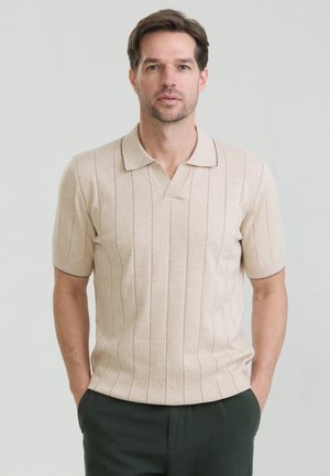 Man met bruin haar die een beige, gebreide poloshirt met korte mouwen en verticale strepen draagt, staand met handen in de zakken van een donkere broek.