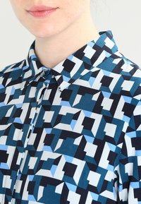 Button-up shirt featuring a geometric pattern in shades of teal, navy, and light blue. Made from a smooth fabric with a collared neckline.