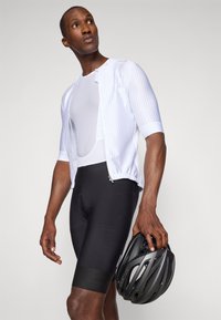 Cyklistický outfit pozostávajúci z bielej rebrovanej bundy na zips, čiernych šortiek a čiernej prilby, ktorý zdôrazňuje elegantný dizajn a textúru materiálu.