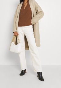 Femme portant un manteau beige, un chemisier marron, un pantalon blanc, des bottes noires, tenant un sac à main blanc avec une anse chaîne dorée, debout devant un fond blanc.