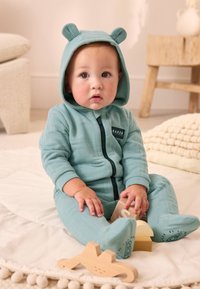 Lysebl å, tekstureret babydragt med hætte og bjørneører, med lynlås foran og logo-patch. Babyen holder træklodser, mens den sidder.