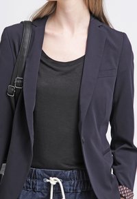 Žena v námořnickém sakě přes černý top a modrých kalhotách se šňůrkou, nese černou koženou tašku přes rameno.