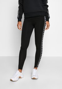 Czarne legginsy o dopasowanym kroju, z białymi paskami po bokach i wzorem logo. Połączone z białymi butami sportowymi dla sportowego wyglądu.