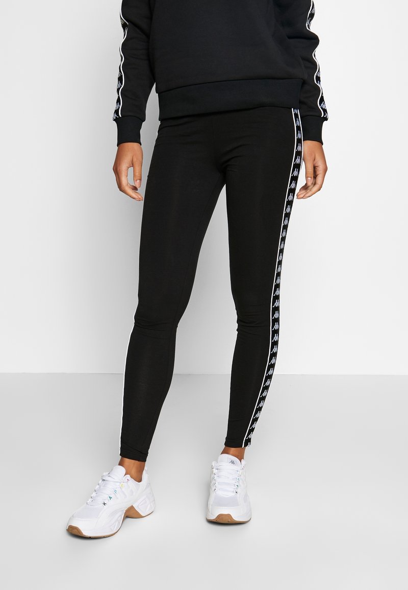 Czarne legginsy o dopasowanym kroju, z białymi paskami po bokach i wzorem logo. Połączone z białymi butami sportowymi dla sportowego wyglądu.