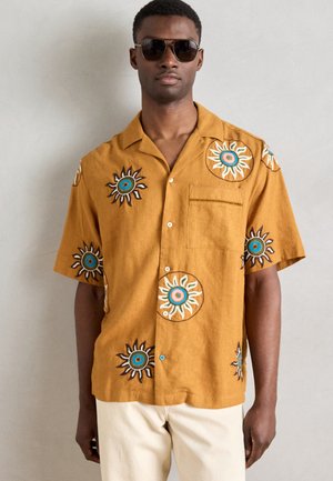 Rövid ujjú, gombos ing mustár sárga anyagból, barna, fehér és kék hímzett napmintákkal. Tartalmaz egy zsebet a mellrészen.