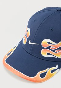 Μπλε σκούφος μπέιζμπολ με κεντημένο σχέδιο φλόγας σε πορτοκαλί, κίτρινο και λευκό. Διαθέτει καμπύλη ρίγα και λογότυπο της Nike σε λευκό χρώμα.