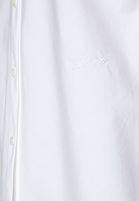 Camicia bianca con bottoni, con un sottile ricamo della scritta "Karl" sul petto, mostrata in primo piano con pieghe visibili e bottoni.