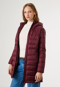 Bordo puffer jakna s kapuljačom, sa quilted teksturom, punim patentnim zatvaračem i džepovima, nosi se preko bijelog pulovera i plavih traperica.